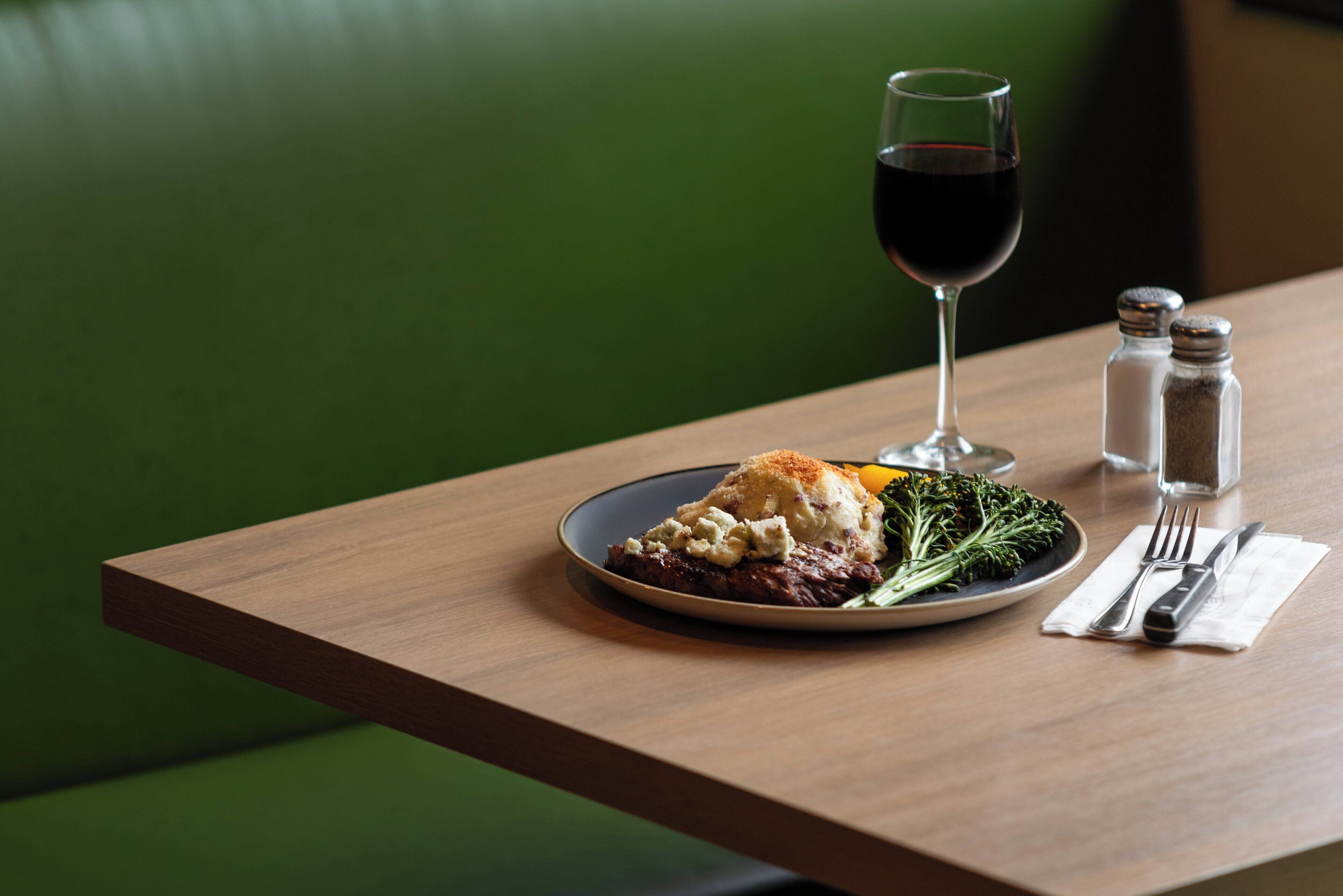 A plate of food with a glass of red wine, salt and pepper shakers, and cutlery on a wooden table.