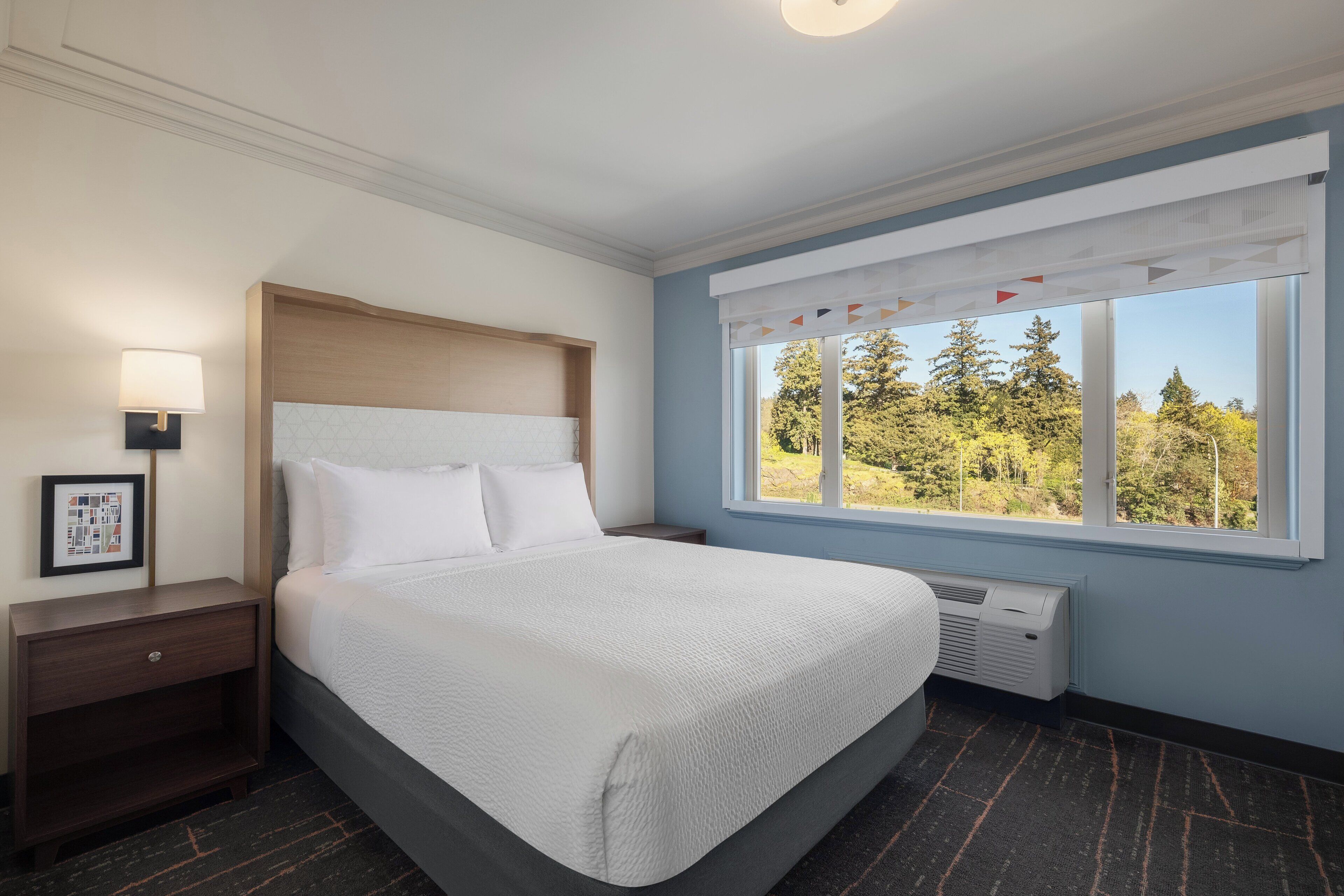 Ein Hotelzimmer mit Bett, Nachttisch, Lampe, Fenster mit Blick auf Bäume und einem gerahmten Bild an der Wand.