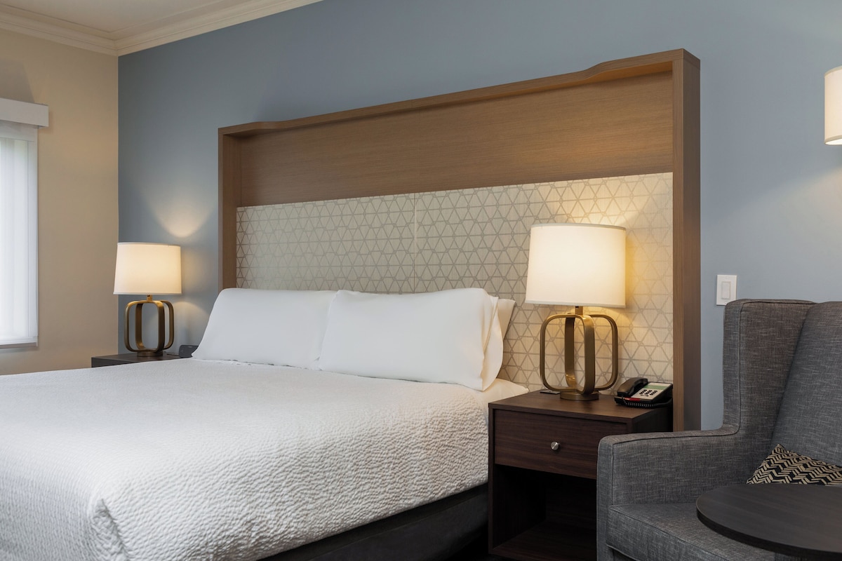 A hotel room with a large bed, two bedside tables with lamps, a chair, and a wooden headboard.