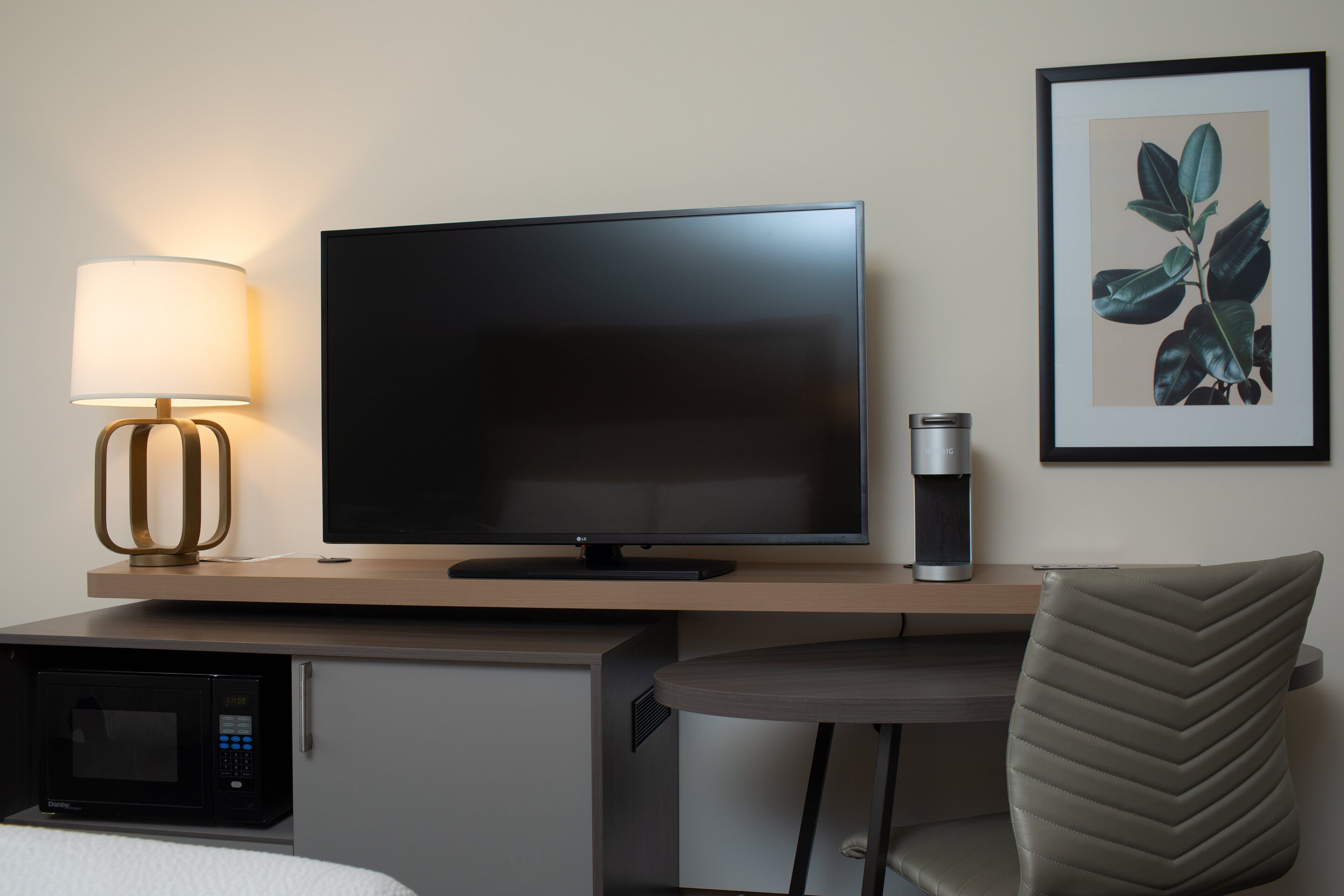 A modern hotel room with a flat-screen TV, a lamp, a framed plant, a coffee machine, a microwave, and a chair.