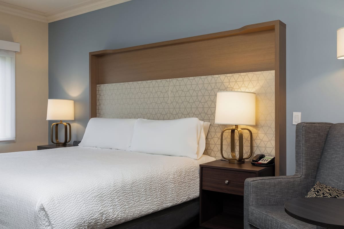 A hotel room with a large bed, two bedside tables with lamps, a chair, and a wooden headboard.