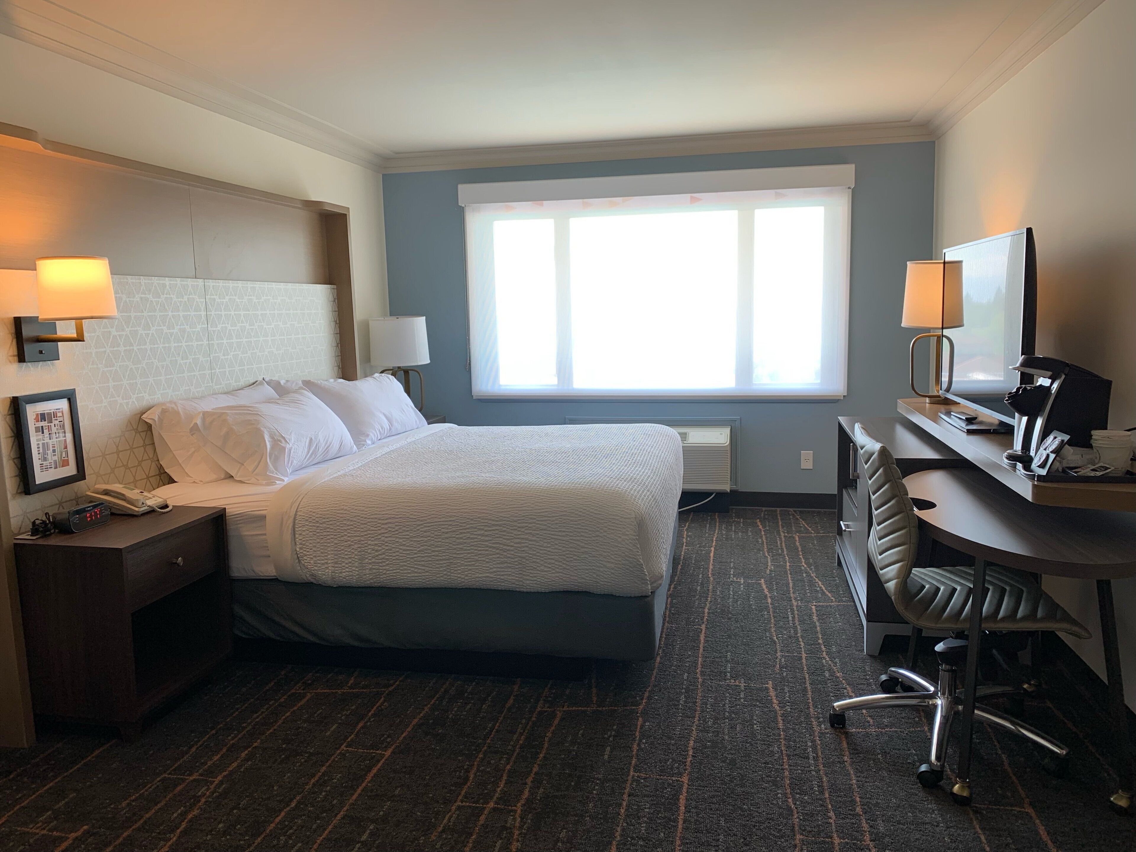 A hotel room with a bed, bedside lamps, a desk with a chair, a window, and a picture frame on the wall.