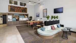 A modern hotel lobby with a seating area, a dining table, and a kitchen in the background.