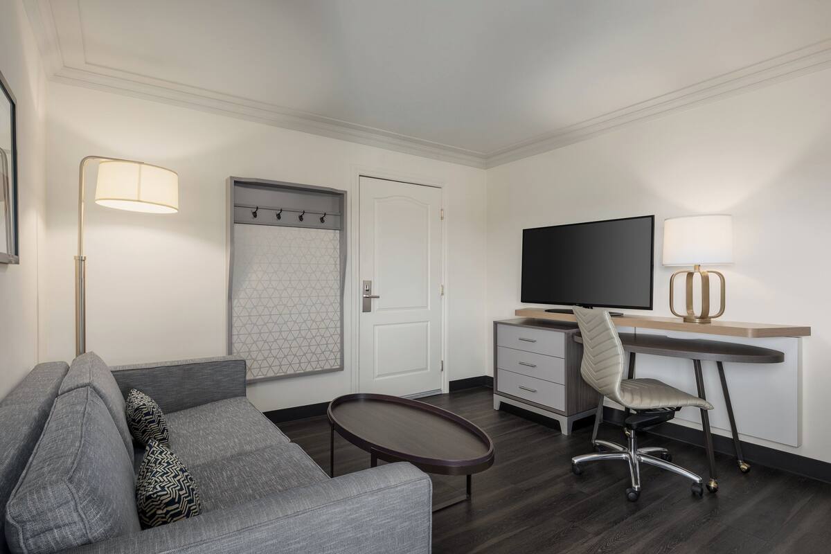 Habitación de hotel moderna con sofá gris, una mesita, un escritorio con silla, televisor de pantalla plana y una lámpara.