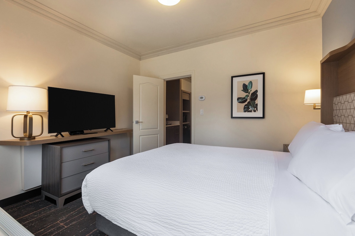 A hotel room with a bed, a television on a stand, a desk with drawers, a door to a closet, and a framed picture on the wall.