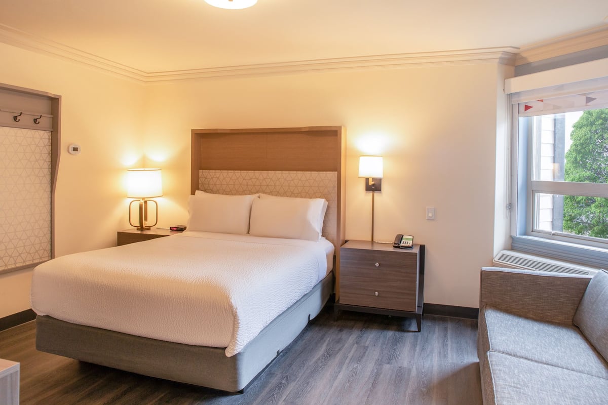 A hotel room with a large bed, bedside lamps, a nightstand, a chair, and a window with a view.