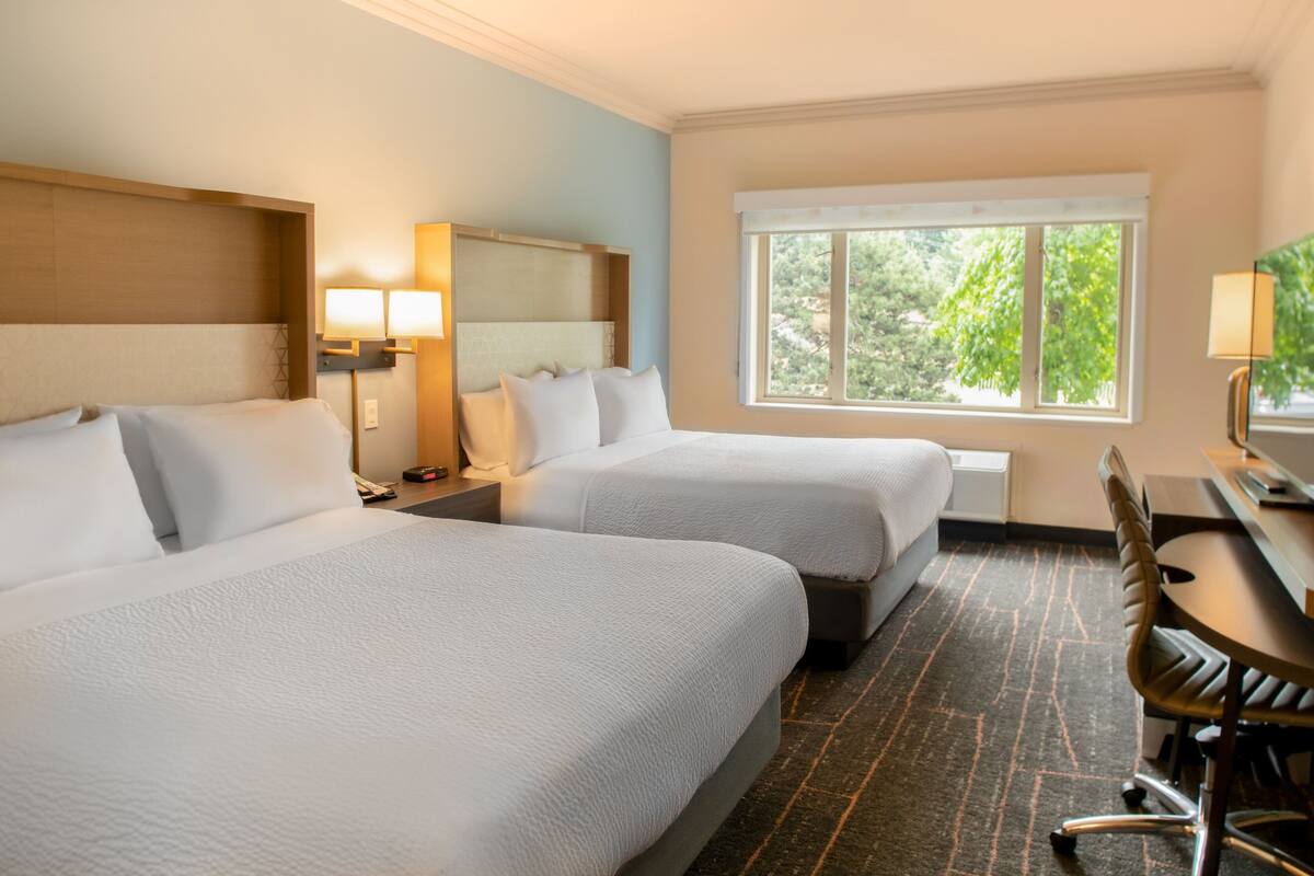 A hotel room with two beds, a desk, and a window with a view of trees.