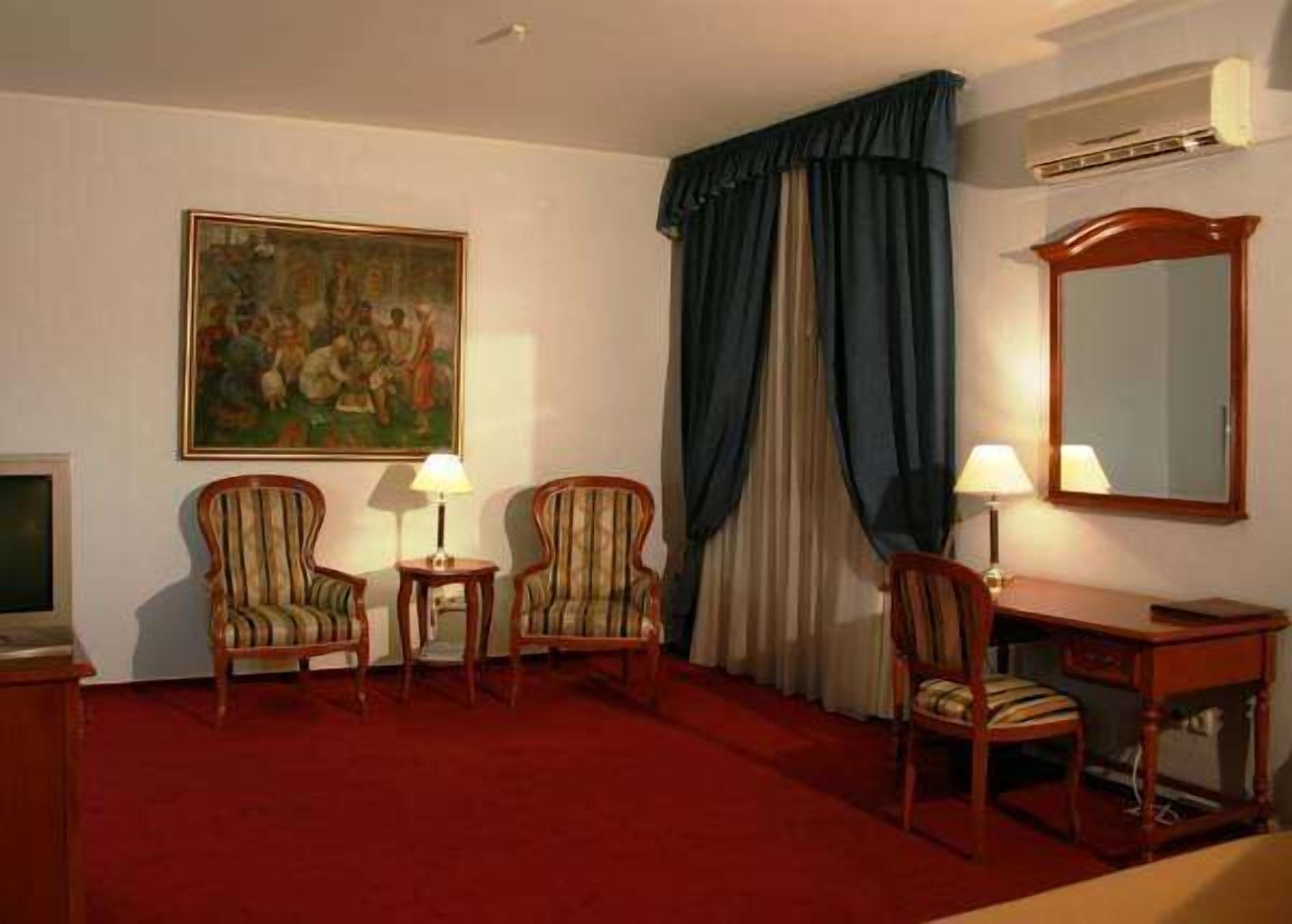 Habitación de hotel con alfombra roja, dos sillones a rayas, una mesita con lámpara, un escritorio con silla, un cuadro grande, una ventana con cortinas azules y un espejo en la pared.