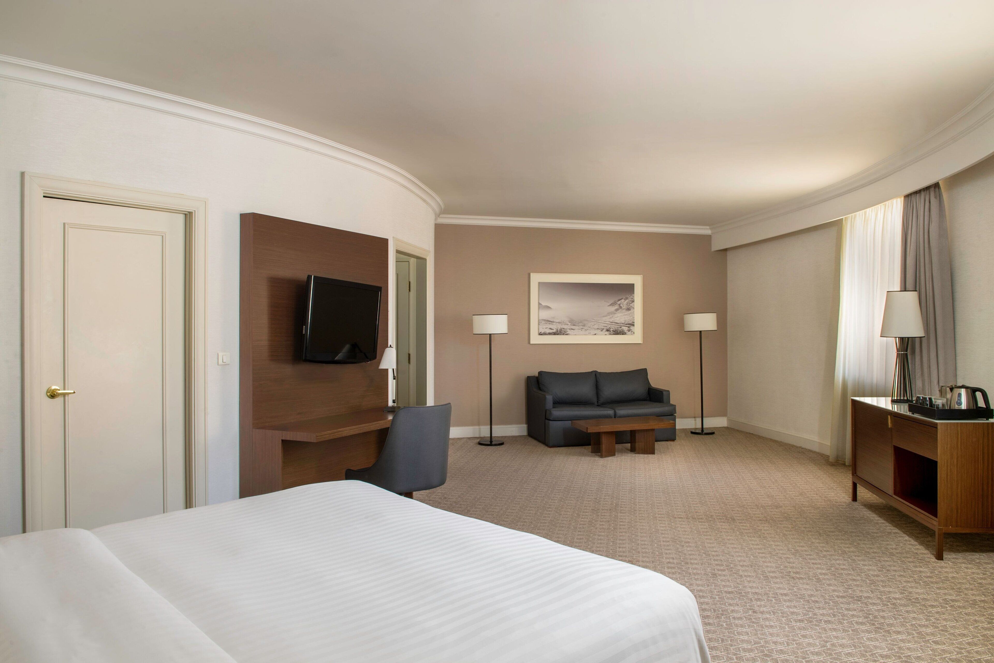 A hotel room with a large bed, a desk with a flat-screen TV, a sofa, two floor lamps, and a painting on the wall.