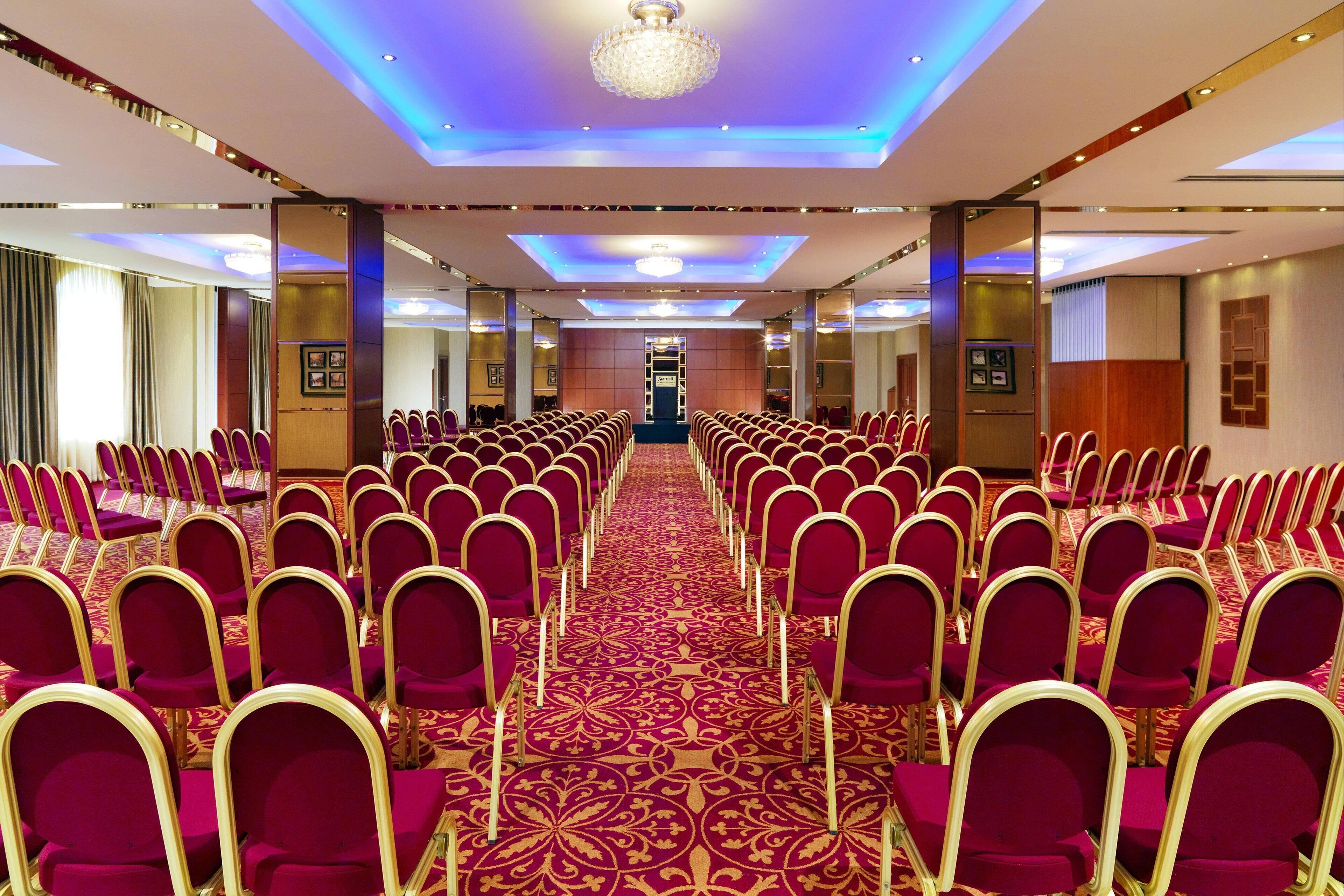 Una sala conferenze con file di sedie rosse, una moquette decorata e un lampadario.