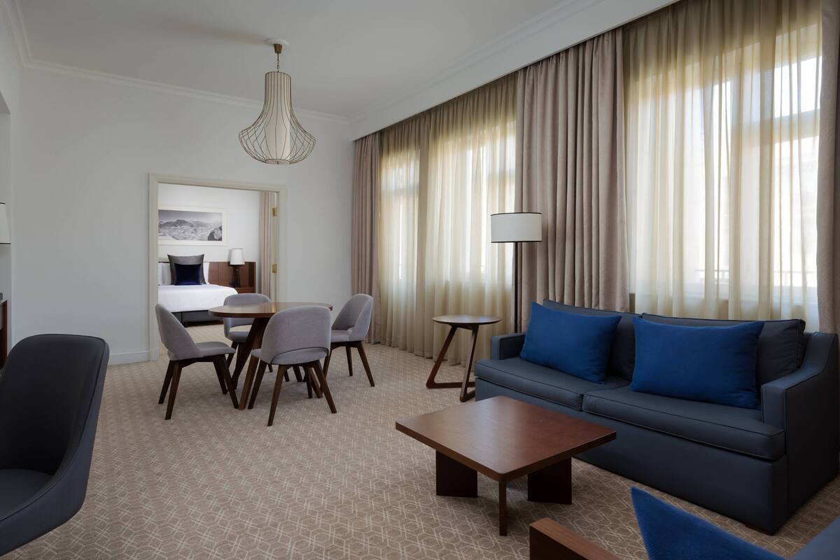 Ein Hotelzimmer mit Sofa, Sesseln, einem Couchtisch, einer Lampe und einem Bett mit Blick ins Freie.