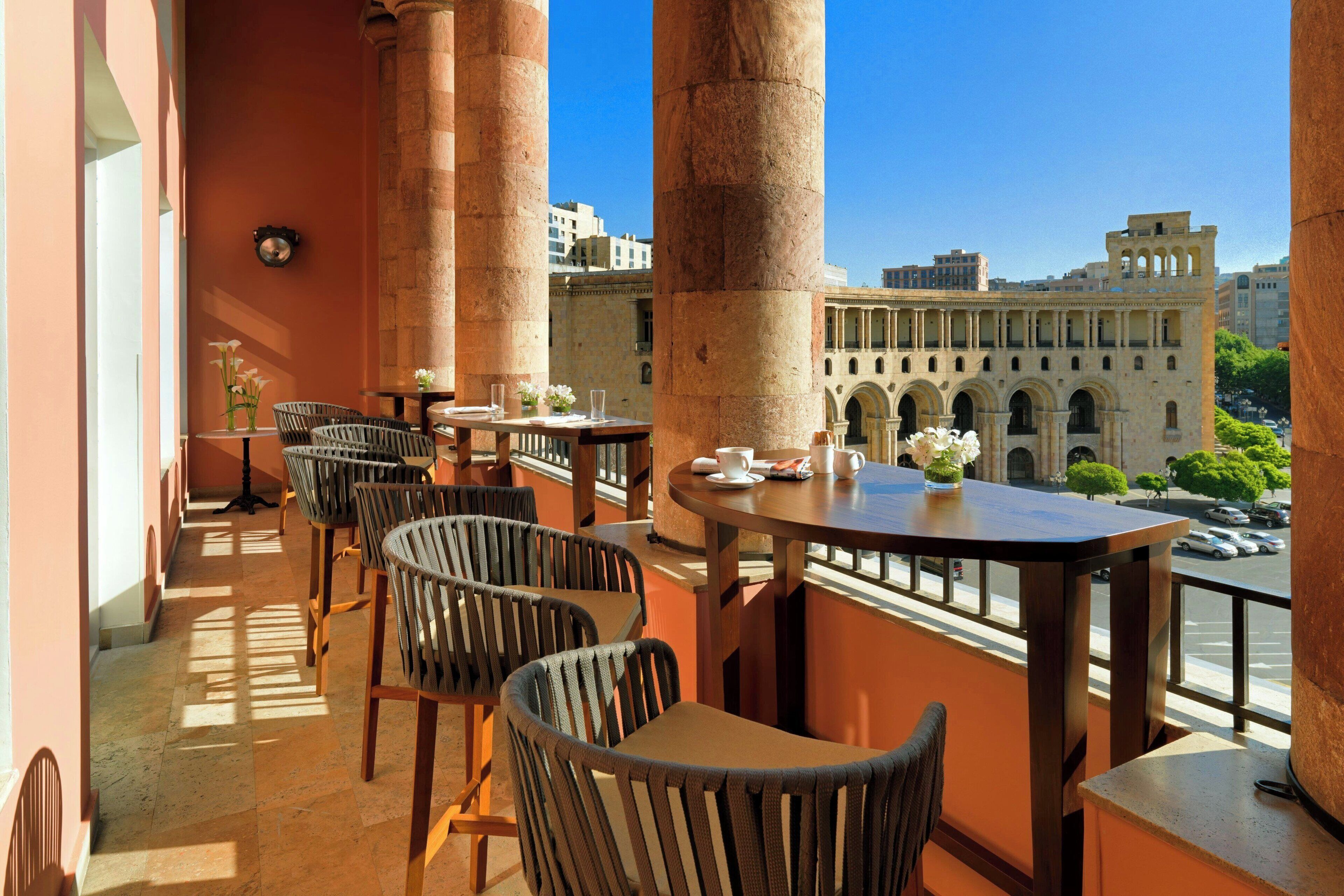 Een balkon met rieten stoelen en tafels, met uitzicht op een stadsgezicht met historische architectuur.