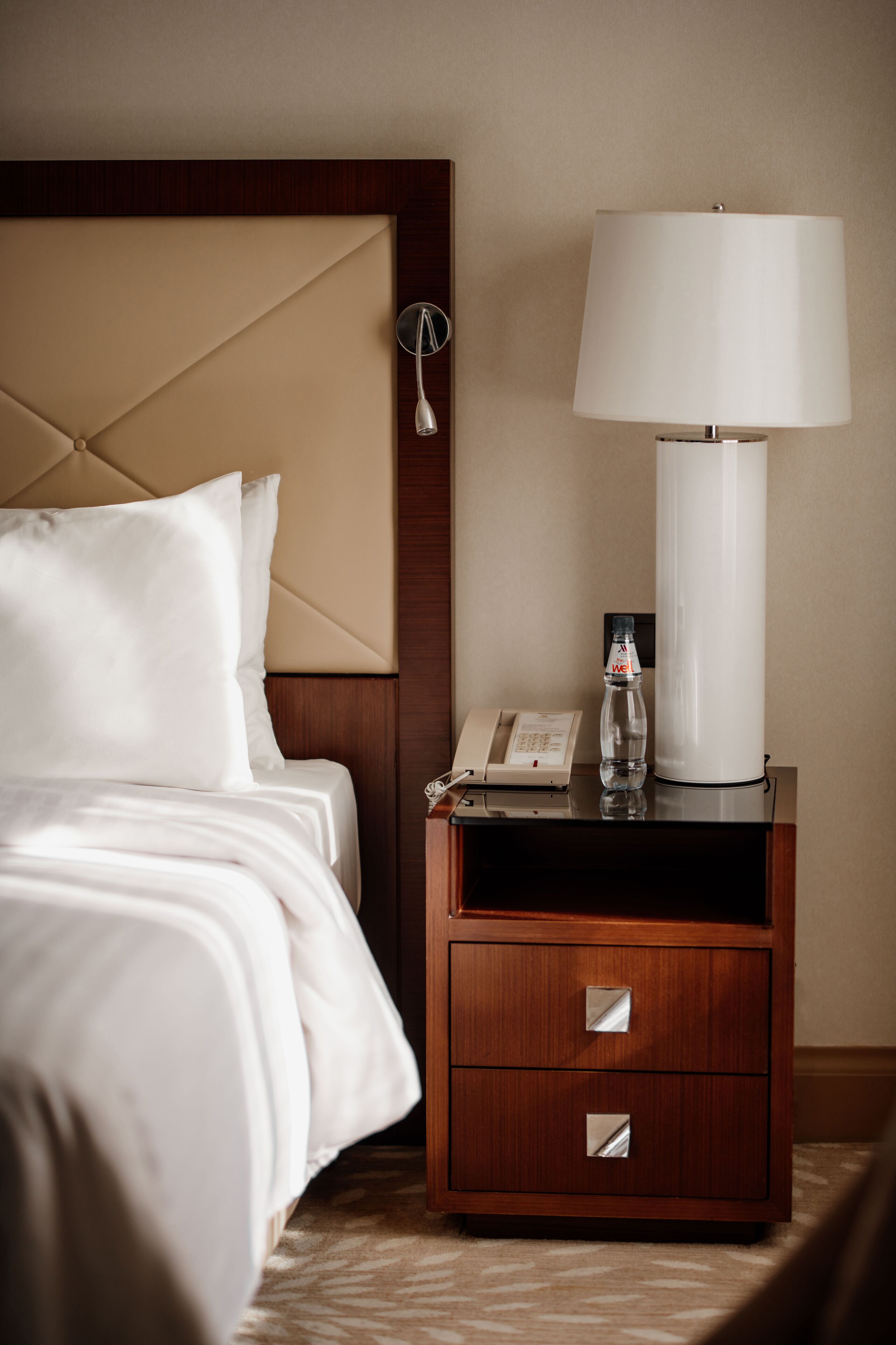 Een hotelkamer met een bed, een houten nachtkastje met een lamp, een telefoon en een fles.