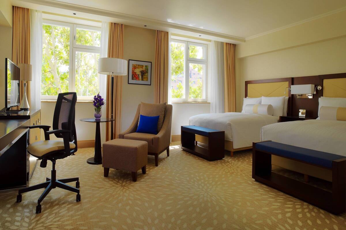 Ein Hotelzimmer mit Bett, Schreibtisch, Stuhl, Hocker und Blick auf Bäume durch das Fenster.