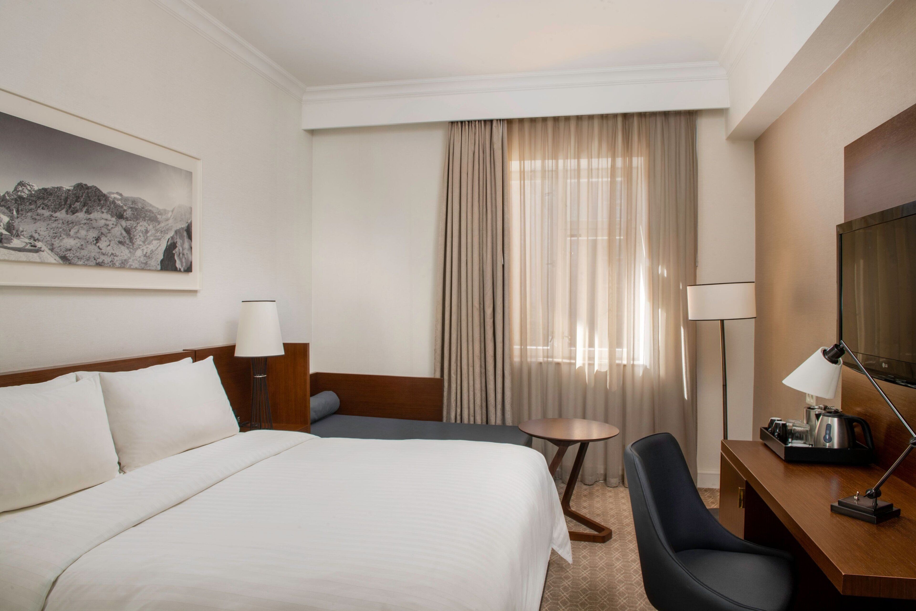 A hotel room with a large bed, a desk with a chair, a television, and a window with curtains.