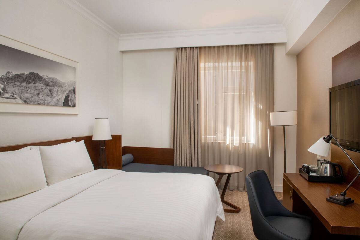 Ein Hotelzimmer mit einem großen Bett, einem Schreibtisch mit Stuhl, einem Fernseher und einem Fenster mit Vorhängen.