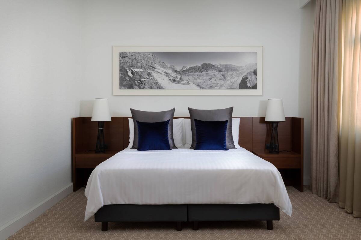 Ein Hotelzimmer mit einem großen Bett, zwei Nachttischen mit Lampen, einem gerahmten Bild an der Wand und einem Fenster mit Vorhängen.