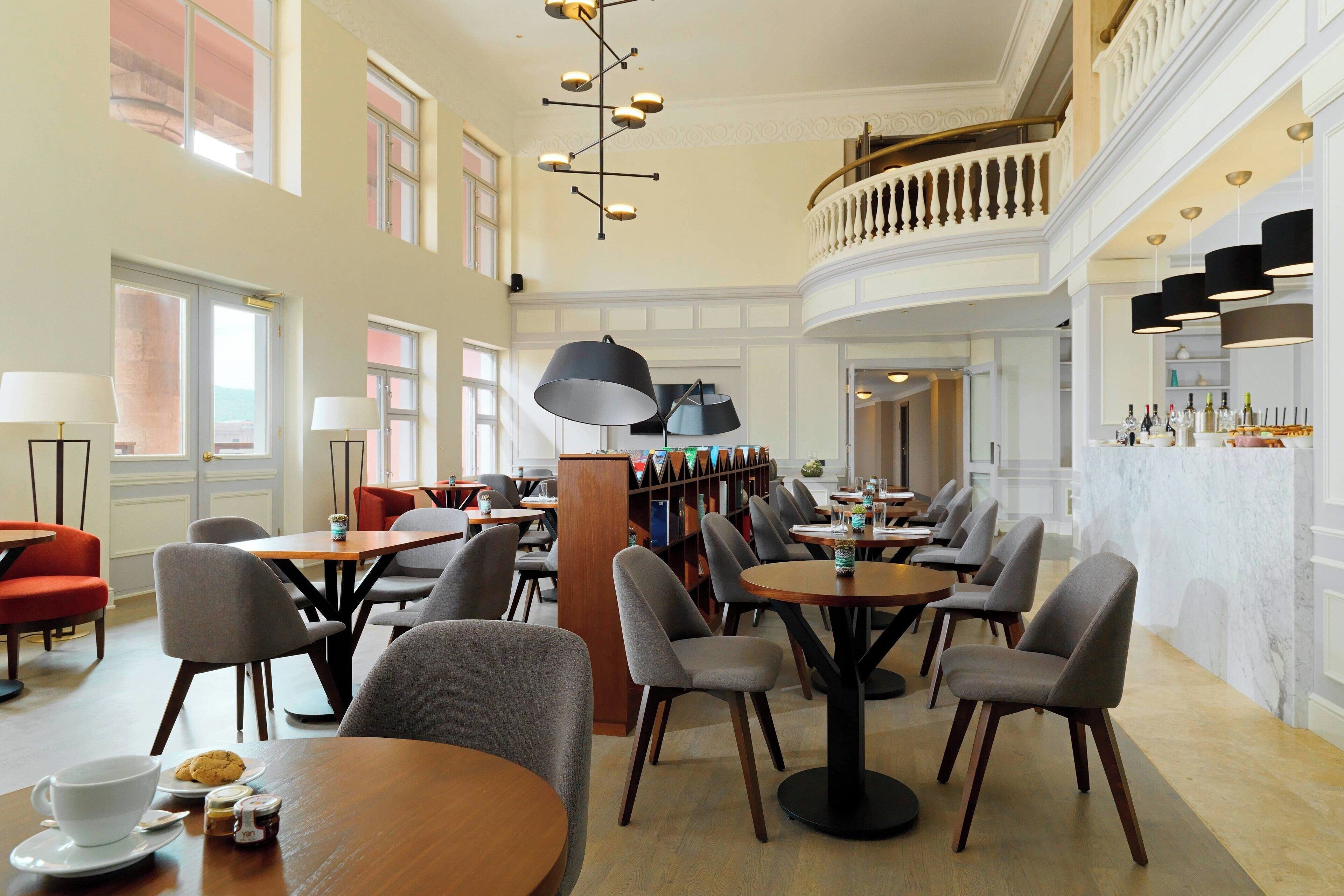A spacious library with multiple tables and chairs, a large wooden bookshelf, and a balcony with a railing.