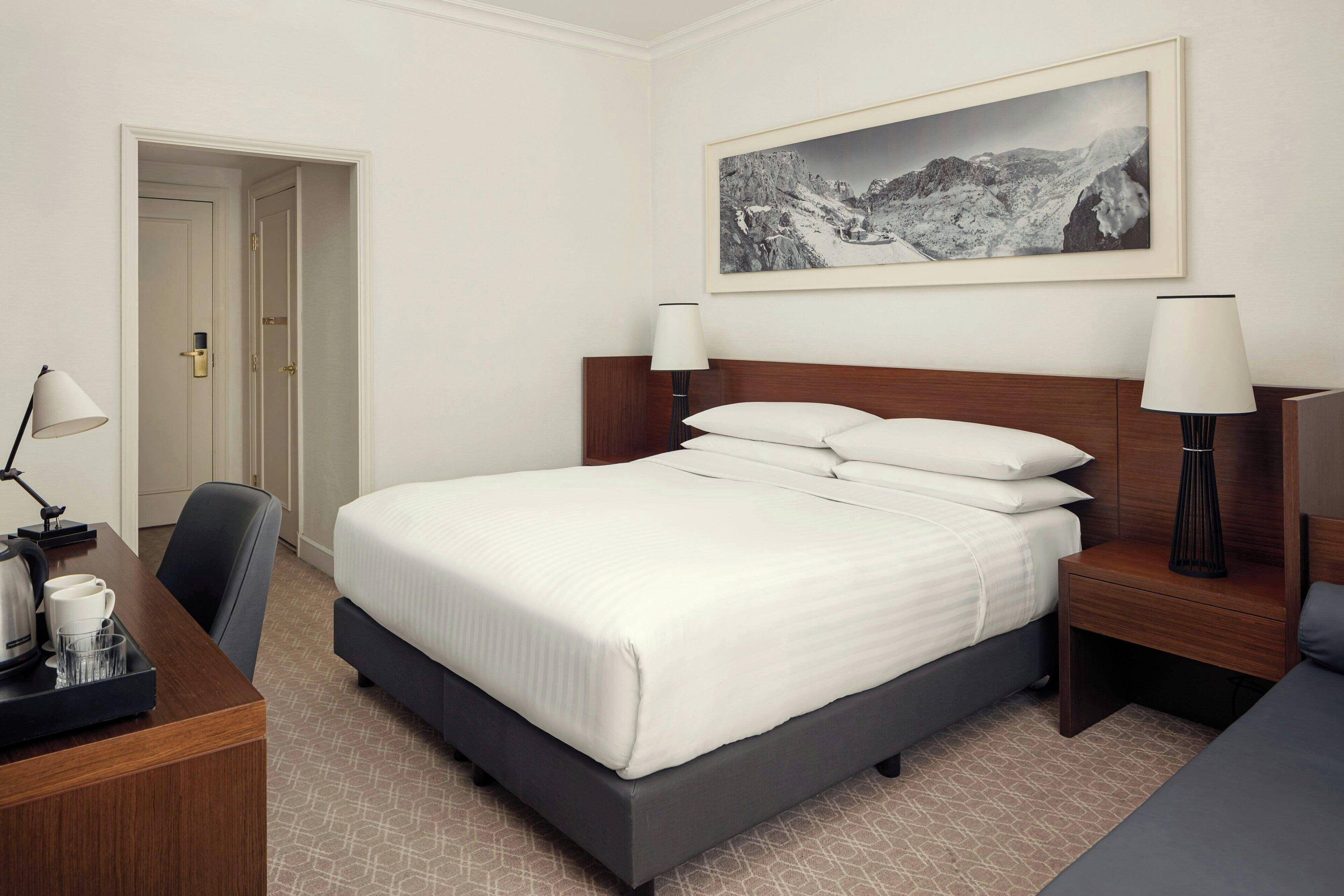 A hotel room with a bed, bedside lamps, a desk with a coffee maker, and a framed picture on the wall.