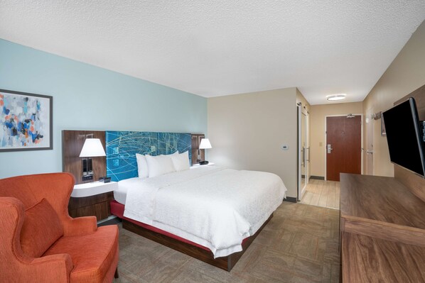 A hotel room with a bed, a red armchair, a desk, a TV, and a painting on the wall. - Hampton Inn & Suites by Hilton West Little Rock (Little Rock)