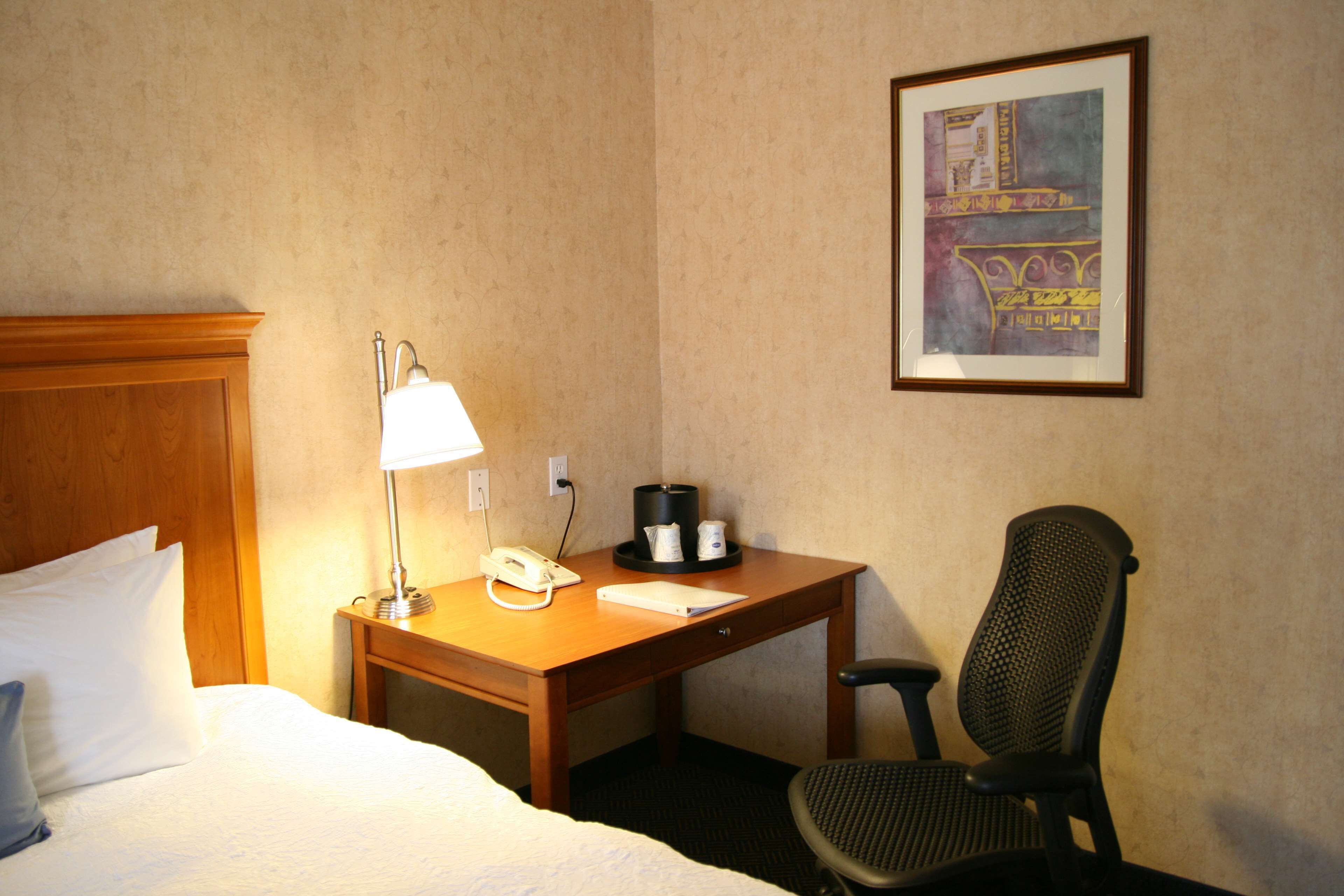 Una habitación de hotel con cama, escritorio con lámpara, teléfono, cafetera y una silla.