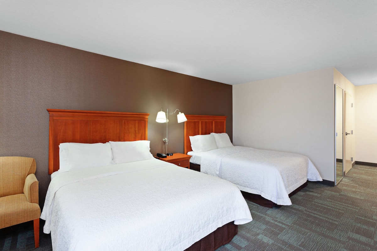 Habitación de hotel con dos camas, cabecera de madera, mesita de noche con lámpara y una silla.