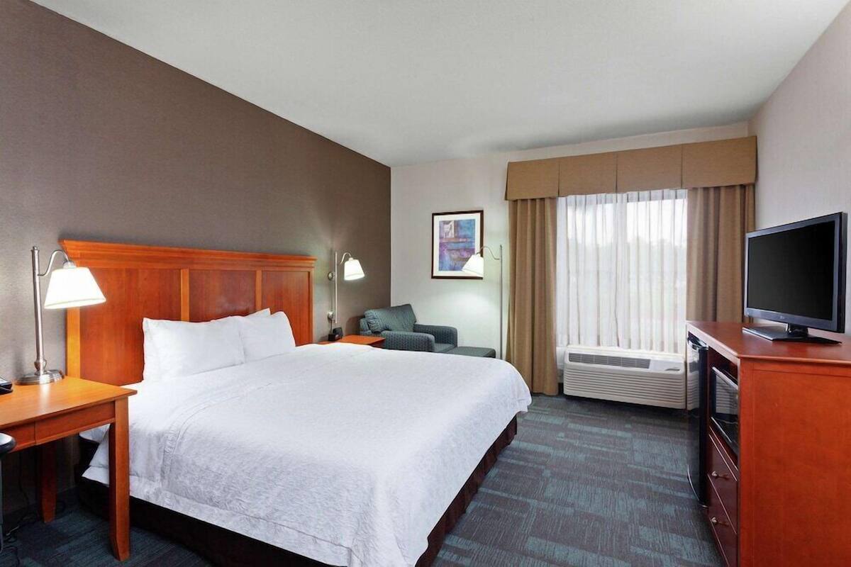 Habitación de hotel con una cama grande, un escritorio con lámpara, televisión, aire acondicionado y una ventana con cortinas.