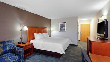 Quarto de hotel com cama, escrivaninha, televisão, abajur e cadeira.