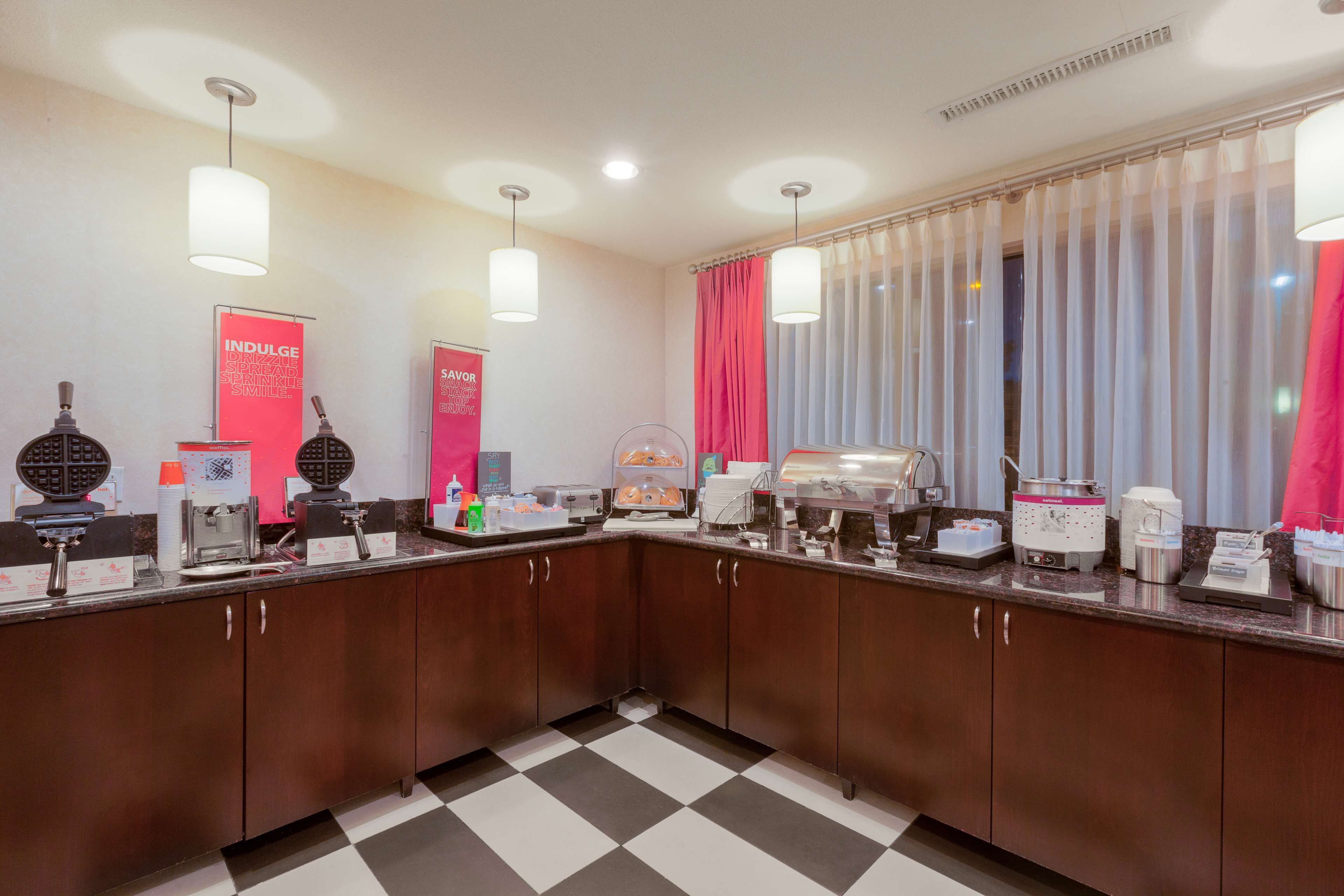 A modern kitchen with a checkered floor, equipped with waffle makers, coffee machines, and a self-service station.
