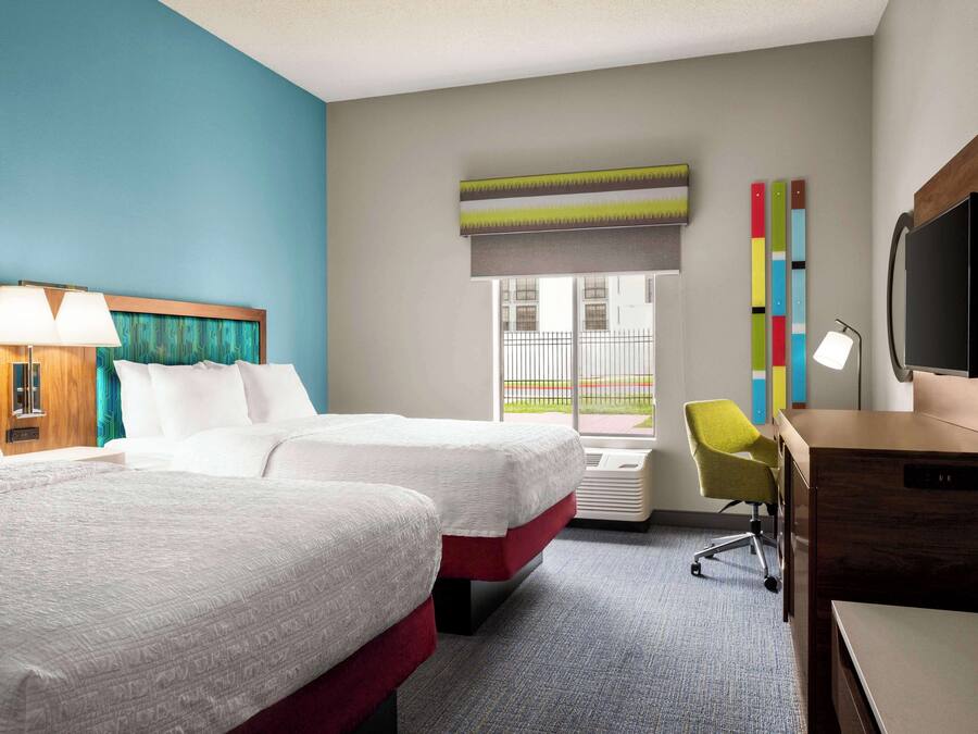 A hotel room with two beds, a desk, a chair, a TV, and a window with a view of the outside.