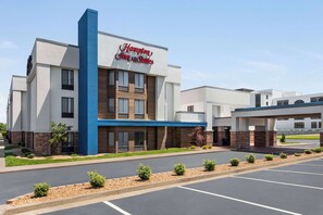 A Hampton Inn & Suites hotel with a modern design, featuring a prominent blue and white color scheme, a parking lot, and a landscaped entrance area.