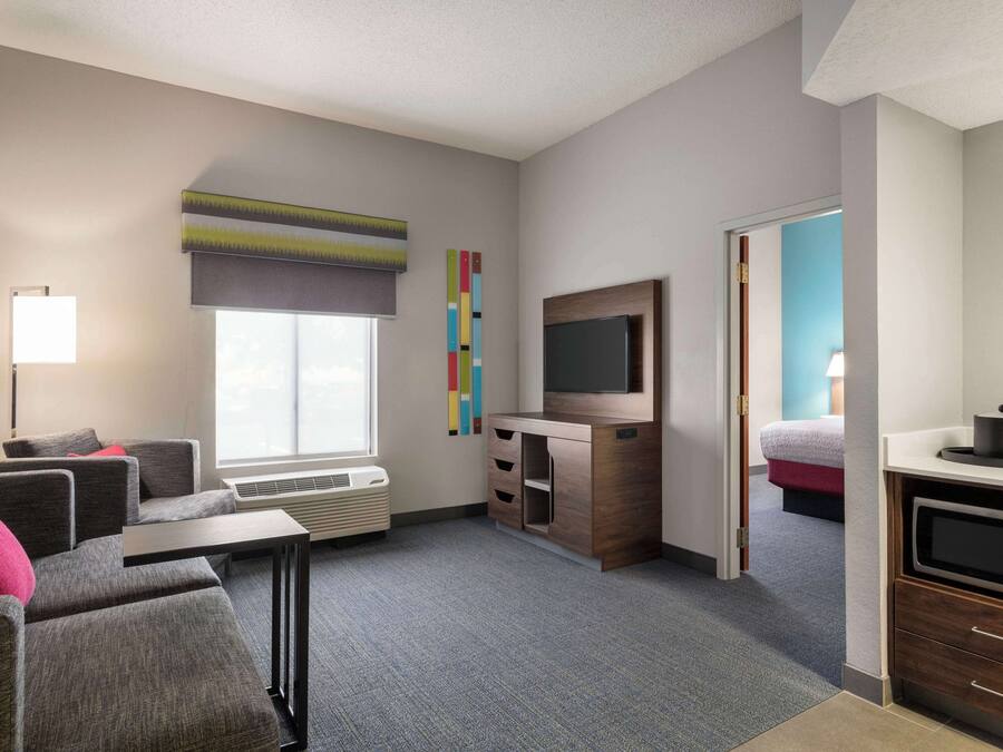 A modern hotel room with a grey sofa, a flat-screen TV, a small kitchenette, and a large window with a view.