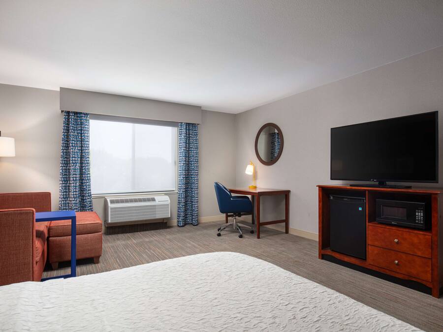 Quarto de hotel com cama, escrivaninha com abajur, televisão, micro-ondas, frigorífico, cadeira e janela com cortinas.