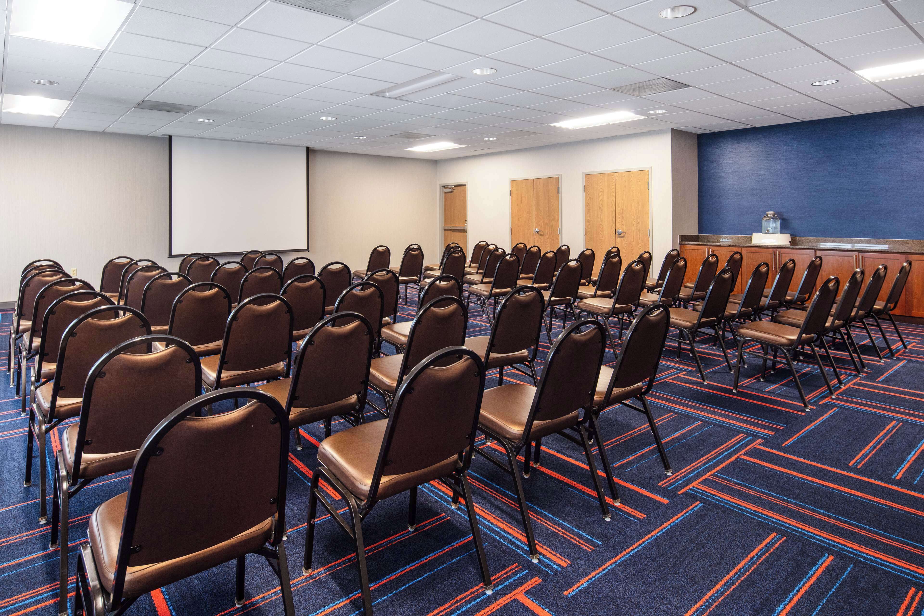 Un salón de conferencias con filas de sillas, una pantalla de proyección y una alfombra azul con líneas rojas.