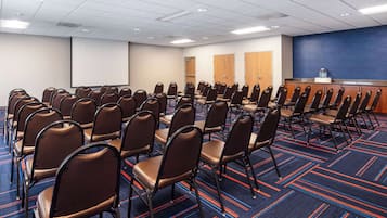 Un salón de conferencias con filas de sillas, una pantalla de proyección y una alfombra azul con líneas rojas.