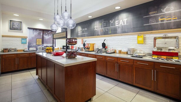 A modern kitchen with wooden cabinets, a central island, and a black backsplash with 'EAT' written on it. - Hampton Inn Medford (Medford)