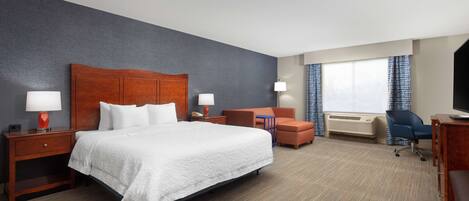 Ein Hotelzimmer mit einem großen Bett, einem Schreibtisch, einem Sessel, einem kleinen Tisch und einem Fenster mit Vorhängen.