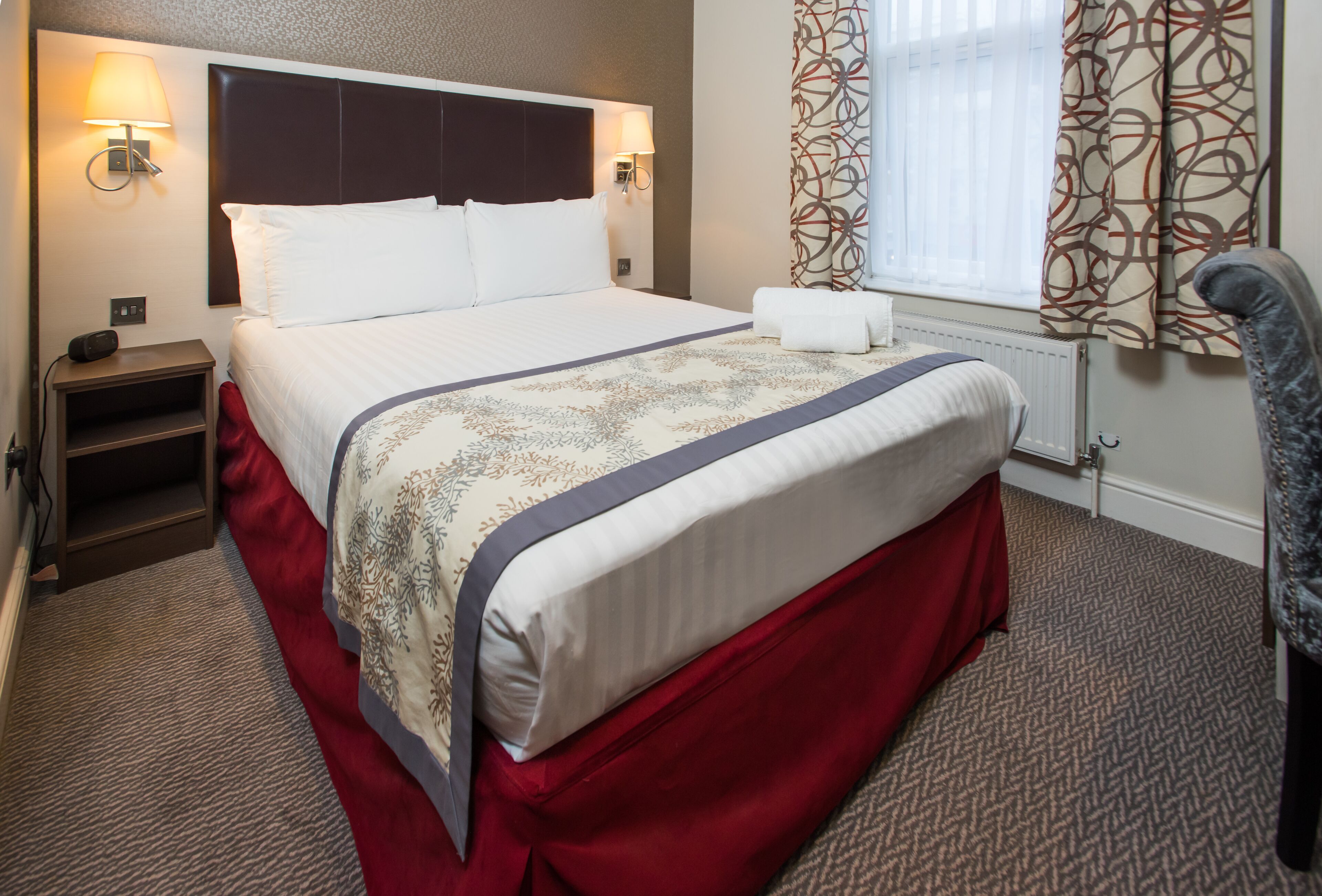 A neatly made bed with a red skirt, a headboard, and two bedside lamps.