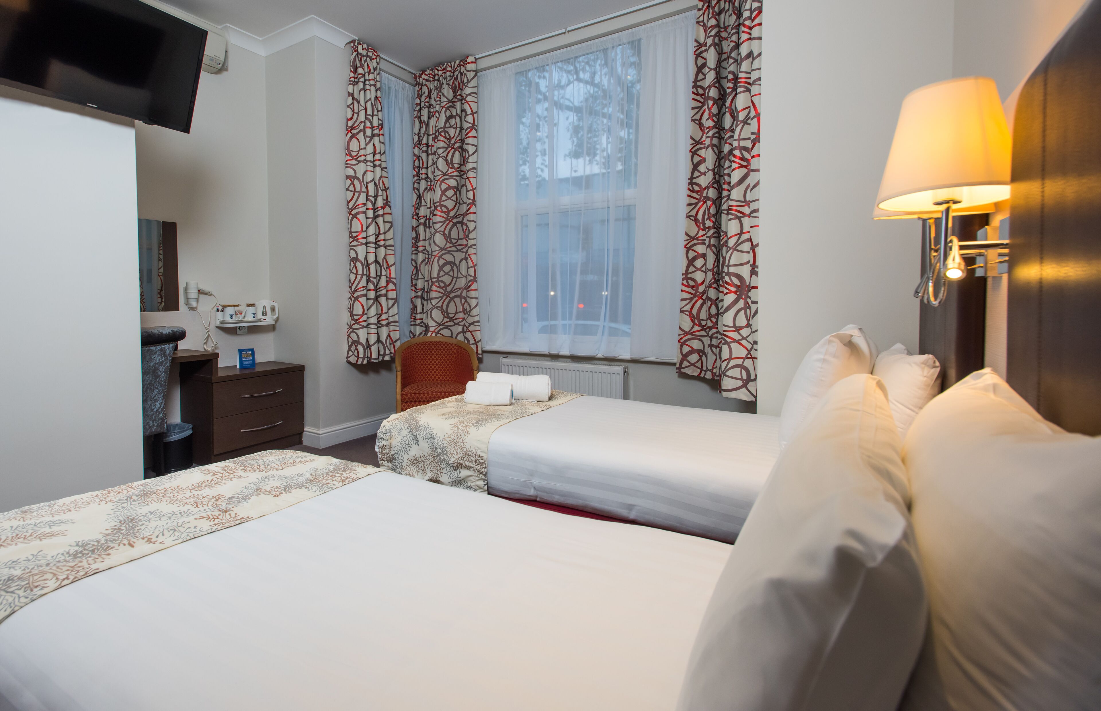 A hotel room with two beds, a TV, a desk with a lamp, and a window with curtains.