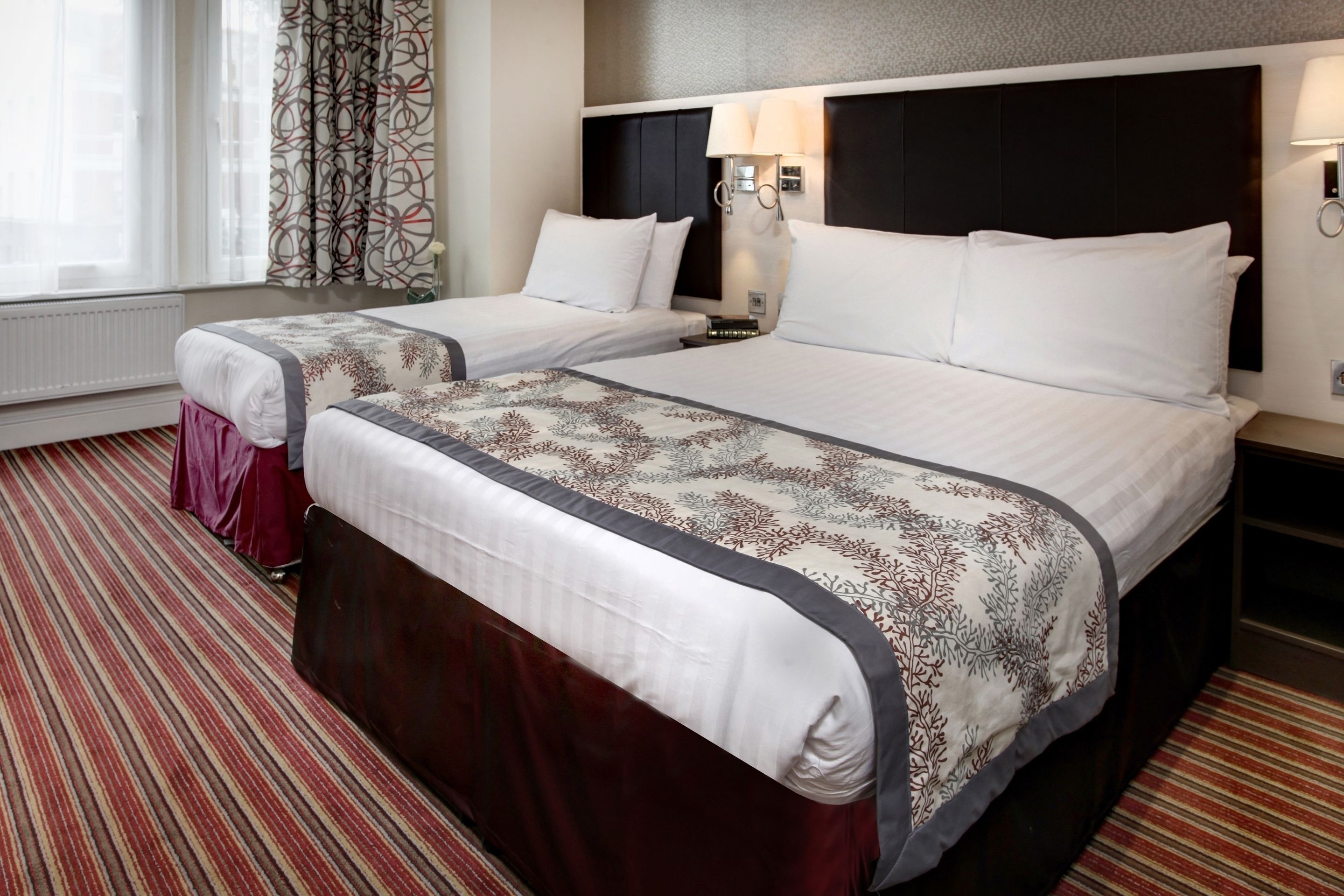 A hotel room with two beds, a large window with curtains, a patterned carpet, and a nightstand with a lamp.