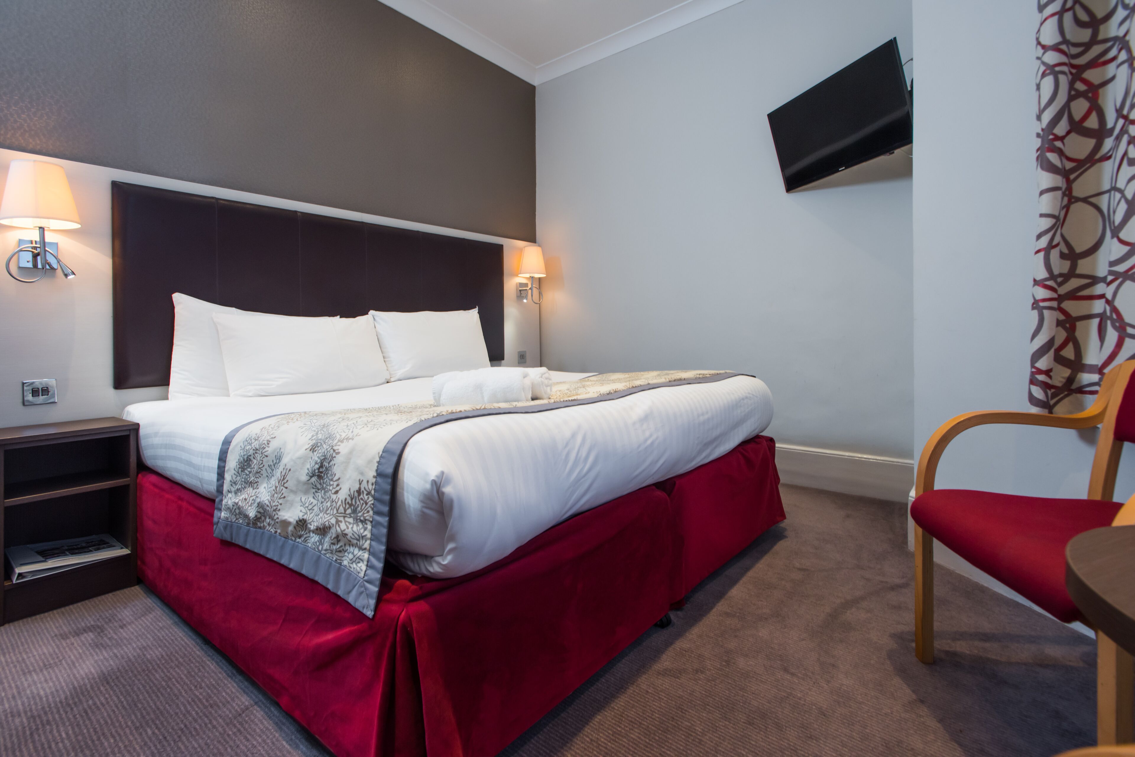 A hotel room with a large bed, a nightstand, a TV mounted on the wall, and a red chair.