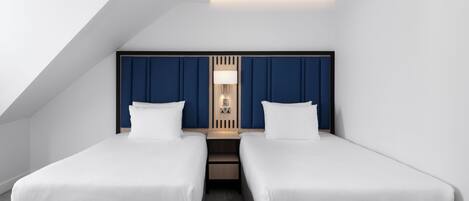 A modern hotel room with a sloped ceiling, two single beds, a nightstand, and a wall-mounted lamp.