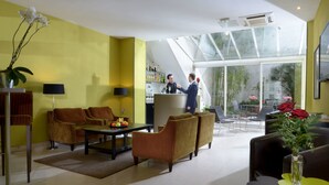 A modern hotel lobby with a yellow wall, a reception desk, seating area, and a large glass window offering a view of the outdoors. - Hôtel Orchidée (Paris)