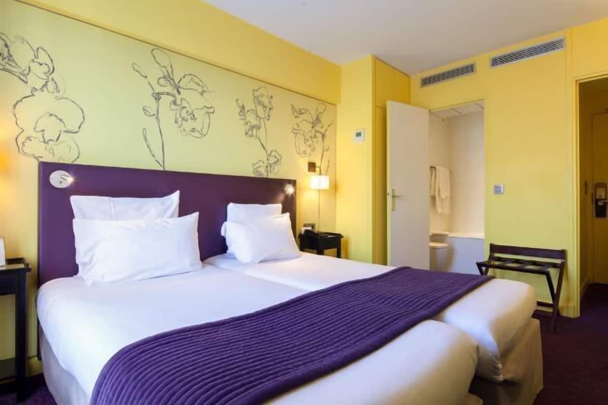 Una cama doble con ropa blanca y un plaid morado, una mesita de noche con lámpara, y una pared con diseños florales.