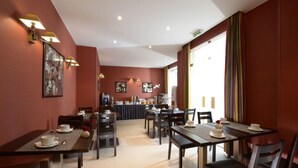 A dining area with tables and chairs, a kitchen in the background, and a large window with curtains. - Hôtel Orchidée (Paris)
