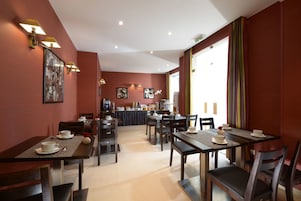 Un área de comedor con mesas y sillas, una cocina al fondo y un ventanal con cortinas.