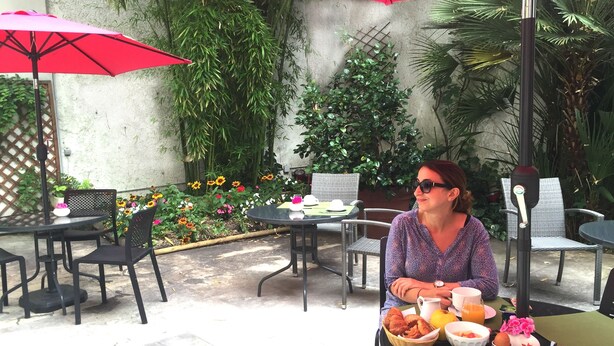 A woman seated at an outdoor table with a meal, surrounded by chairs and tables, with a red umbrella and greenery in the background. - Hôtel Orchidée (Paris)