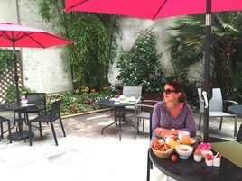 Una mujer sentada en una mesa al aire libre con un plato de comida, rodeada de sillas y mesas, con un paraguas rojo y follaje en el fondo.