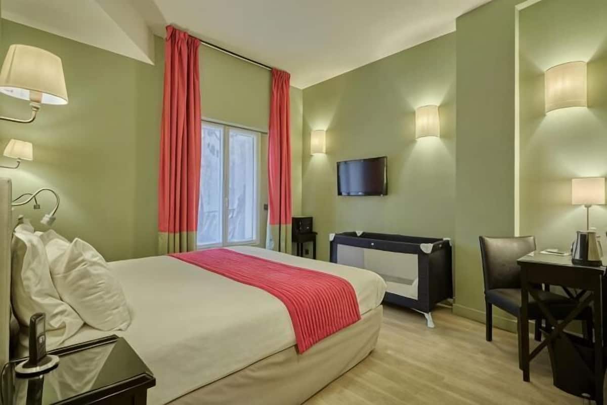 Habitación de hotel con cama, escritorio, silla, televisión y ventana con cortinas.