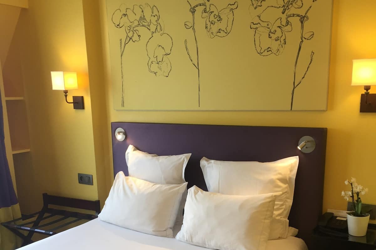 Una cama bien hecha con sábanas blancas y un edredón azul oscuro, cabecera con diseño floral y lámparas de noche sobre una mesita.